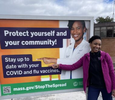 Assoumou-billboard-Twitter Sabrina Assoumou poses in front of and points to the billboard that talks about staying up to date with covid and flu vaccines.