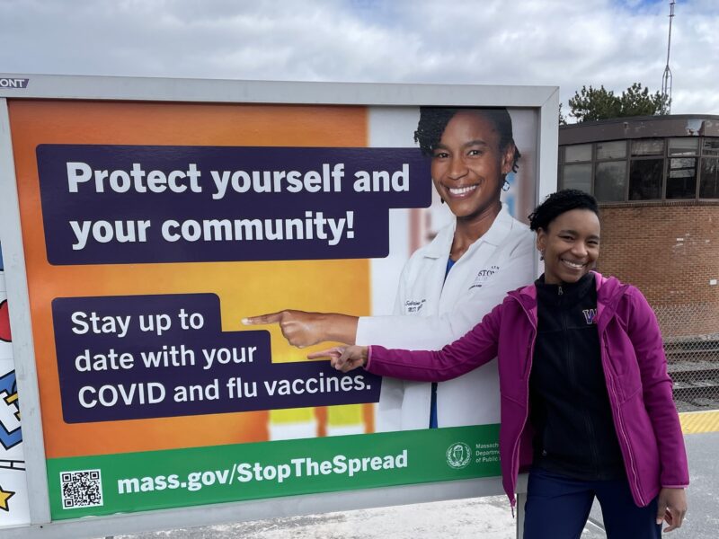 Assoumou-billboard-Twitter Sabrina Assoumou poses in front of and points to the billboard that talks about staying up to date with covid and flu vaccines.