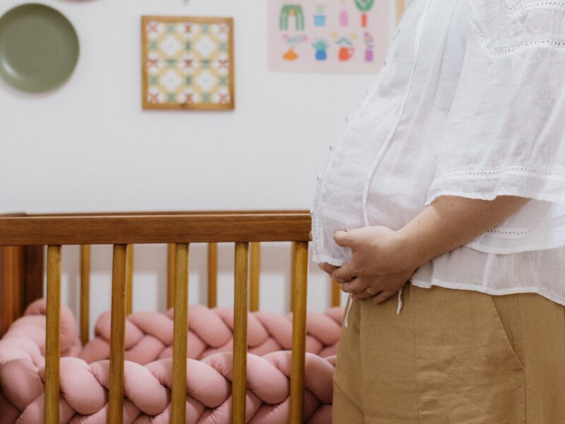 A pregnant woman stands beside a crib.